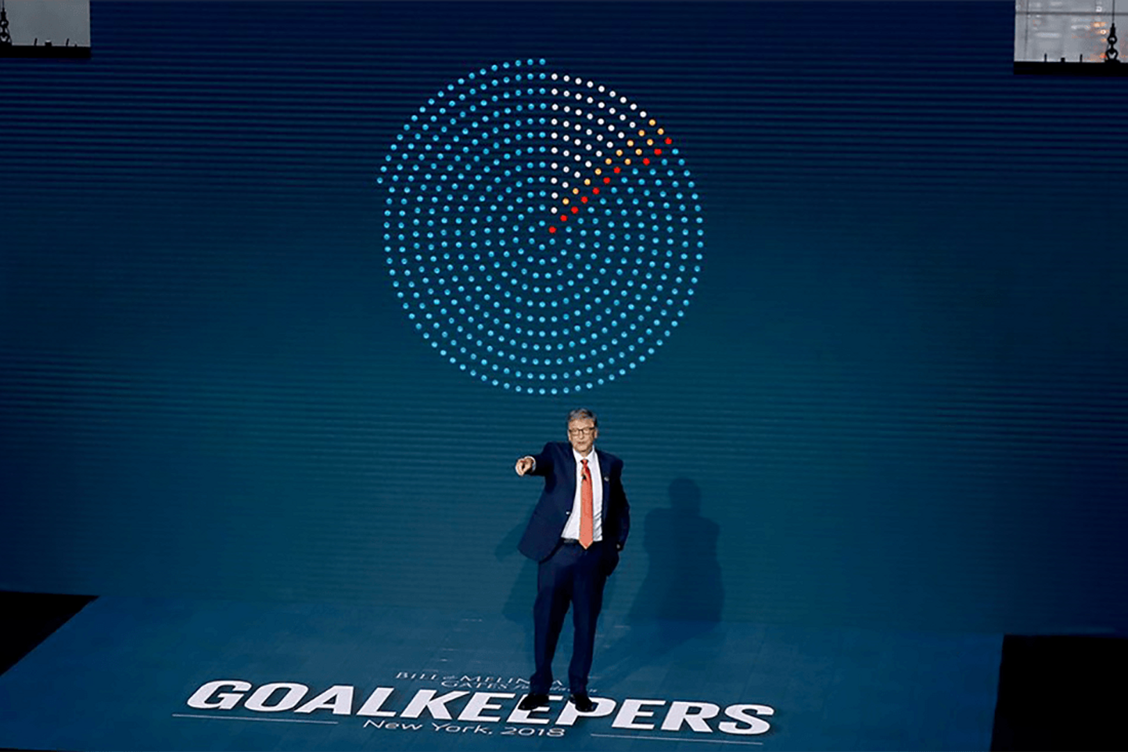 A photograph taken of Bill Gates on stage speaking at a Goalkeepers. Behind him is a pie chart made of dots. Beneath his feet is the word 'Goalkeepers' in larger white letters.