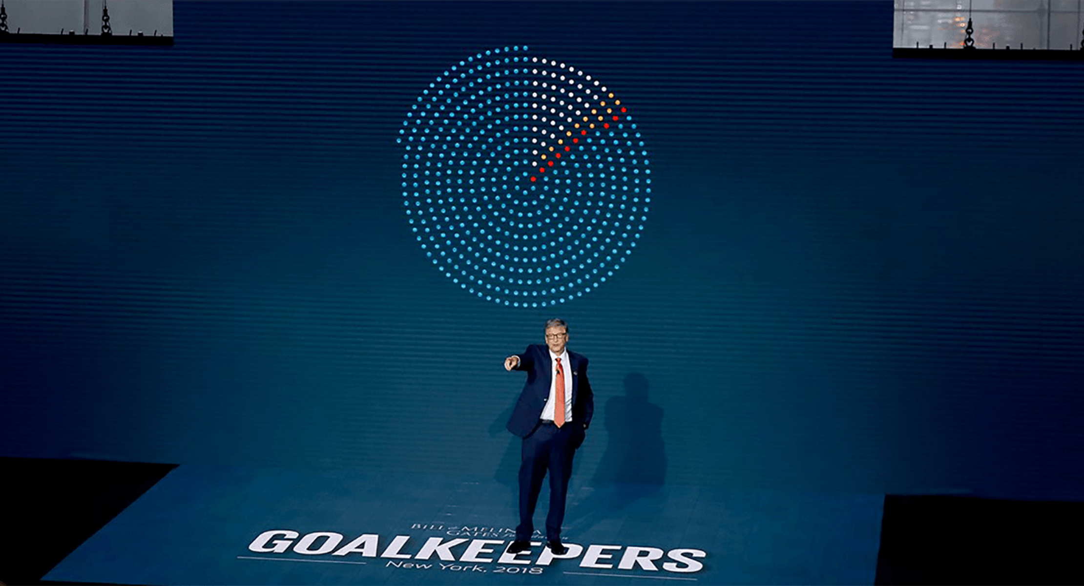 A photograph taken of Bill Gates on stage speaking at a Goalkeepers. Behind him is a pie chart made of dots. Beneath his feet is the word 'Goalkeepers' in larger white letters.