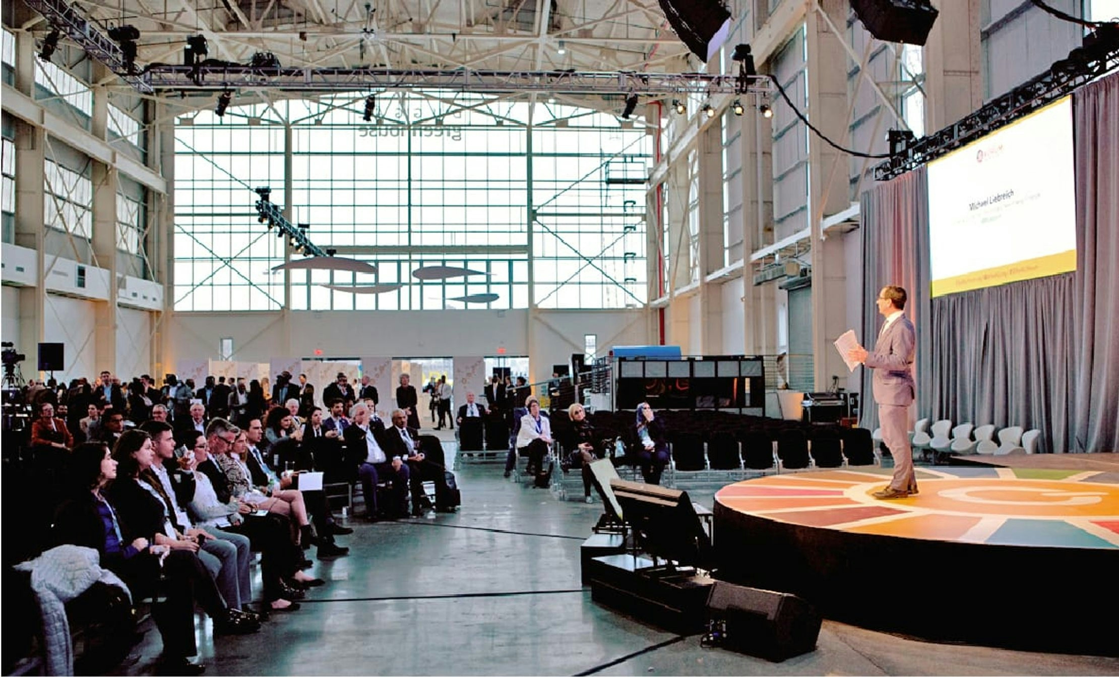A side on shot of a speaker on stage in front of a seated crowd at the Sustainable Energy for All forum. The crowd sit on the left. The speaker on stage is stood on the right of the image.