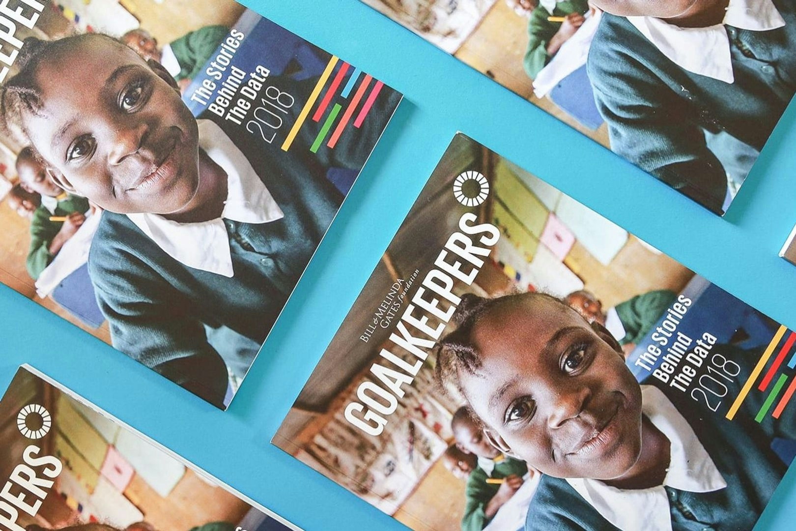 Multiple copies of the Goalkeepers 2018 report laid out side by side over a bright blue background. The cover is titled 'Goalkeepers' with the image of a schoolgirl smiling into the camera.