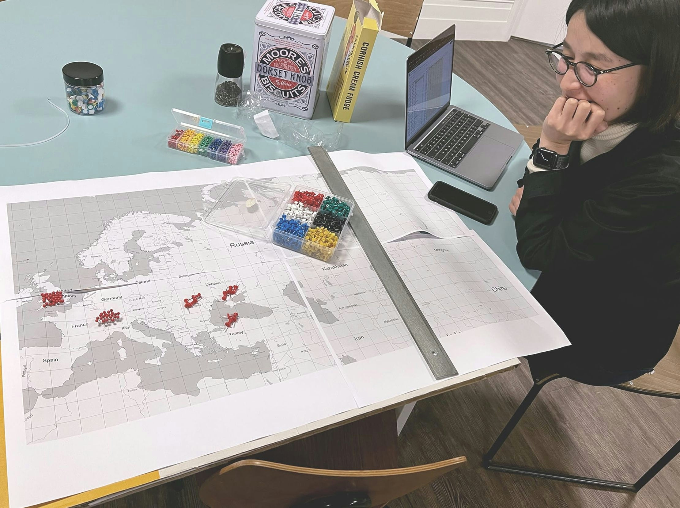 A designer sits at a table with a laptop, paper map of Europe and a box of pins. The map already has several red pins in it as they test fitting all the pins on the map.
