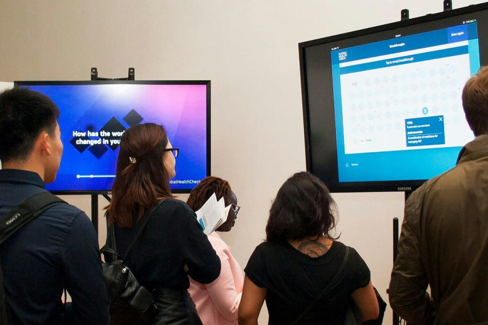 A photo taken during the Global Health Check presentation. It shows a group of people looking at two television screens on stands both displaying different parts of the Global Health Check interactive.