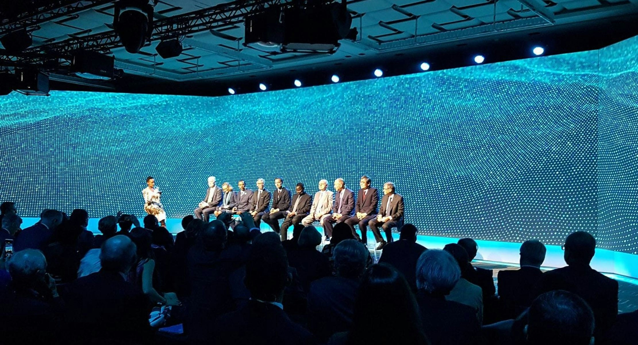 A photo taken during the Cascade presentation. Ten speakers sit on stage in front of three floor to ceiling blue screens that display a mesh moving like the surface of water. In the foreground, an audience sit in the dark watching the stage.