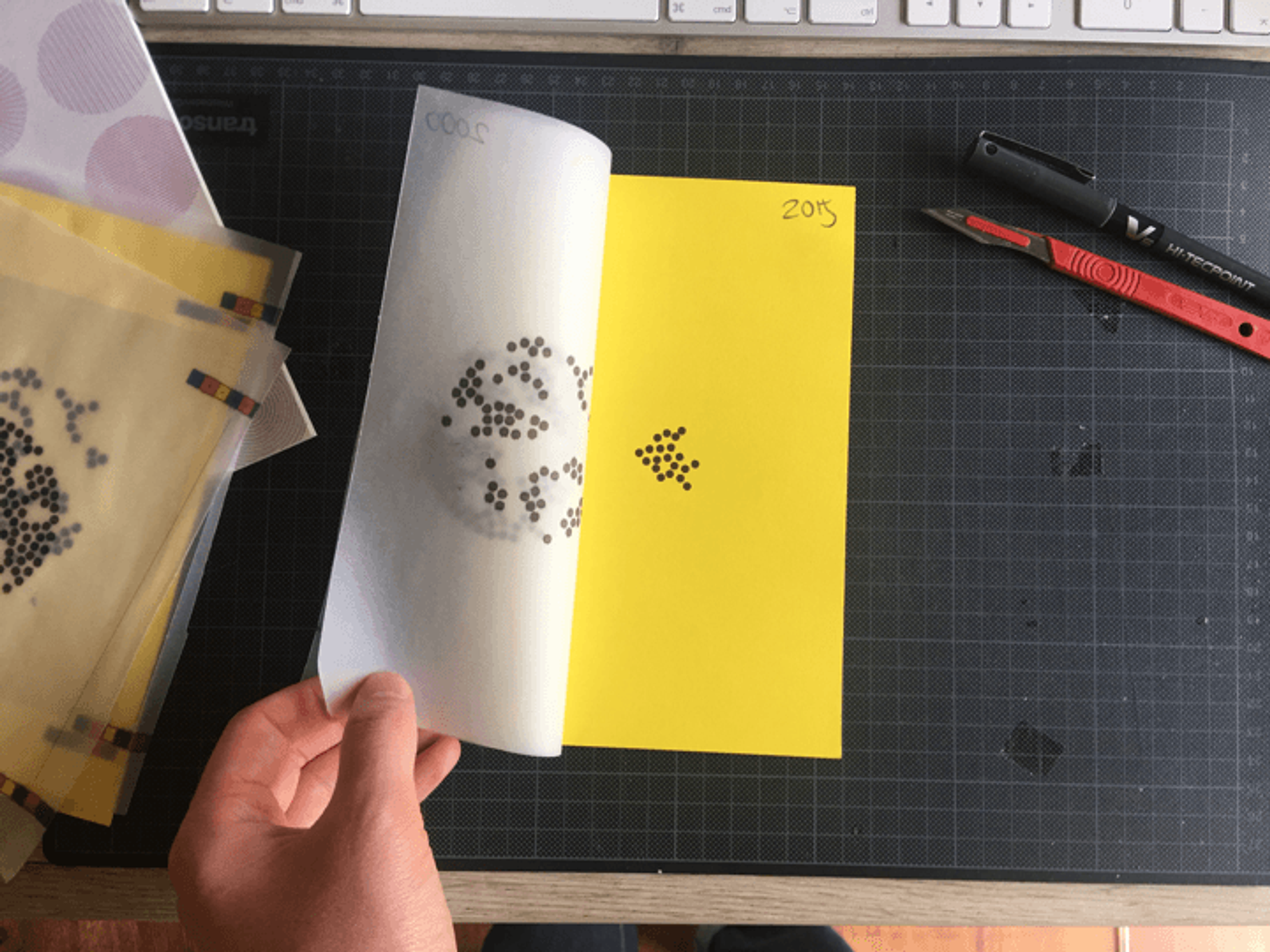 Paper sat on a desk testing transparent paper to layer dots onto a visualization. A hand has half turned the transparent paper open like the page of a book. The page has been turned enough that the yellow paper beneath is clear, it also has a cloud of dots.